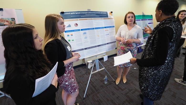One woman asking three female students about their poster displaying their research